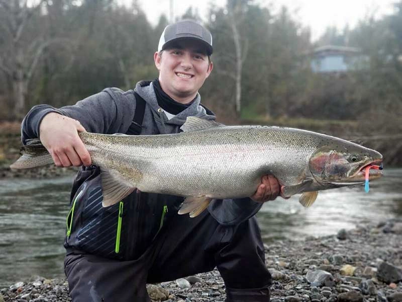 Person on a summer fishing trips in oregon holding a large fish with a lure in its mouth, by a riverbank.